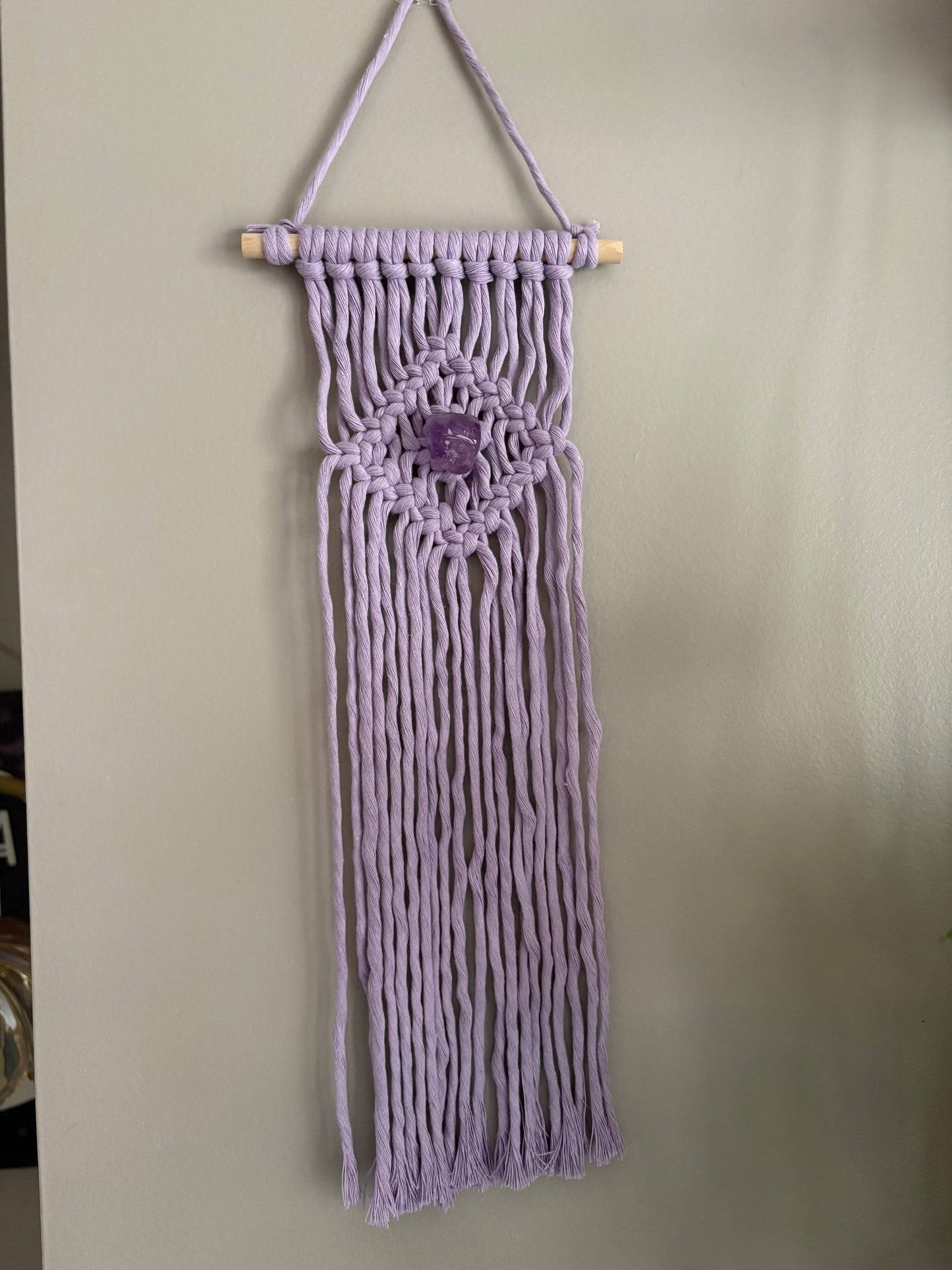 Macrame wall hanging with a central amethyst stone on a plain background