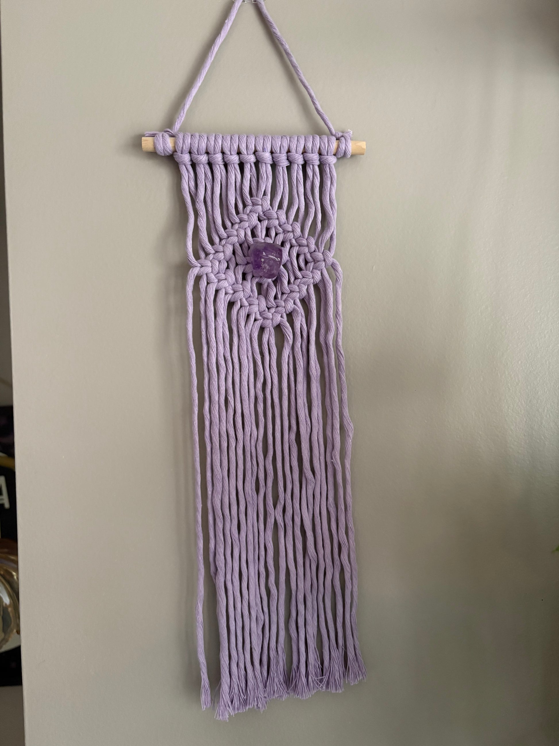 Macrame wall hanging with a central amethyst stone on a plain background